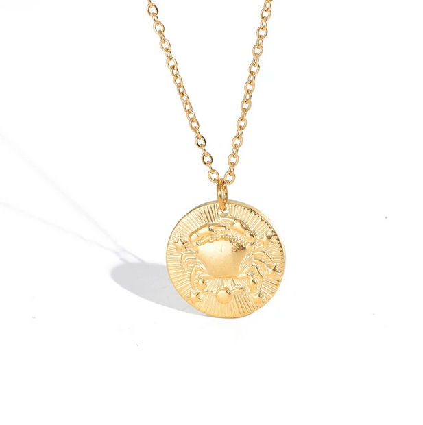 A gold necklace with a dainty chain. The necklace has a coin pendant displaying the astrological sign Cancer of the zodiac.