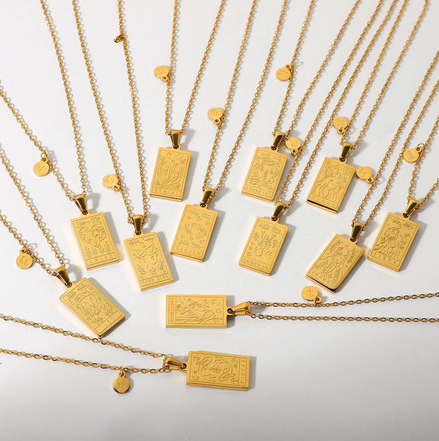 The cardinal necklances laid out on a white table, displaying all styles of the zodiac. The necklaces are a dainty chain with a square style pendant with laser etching to create the artwork of the zodiac. 