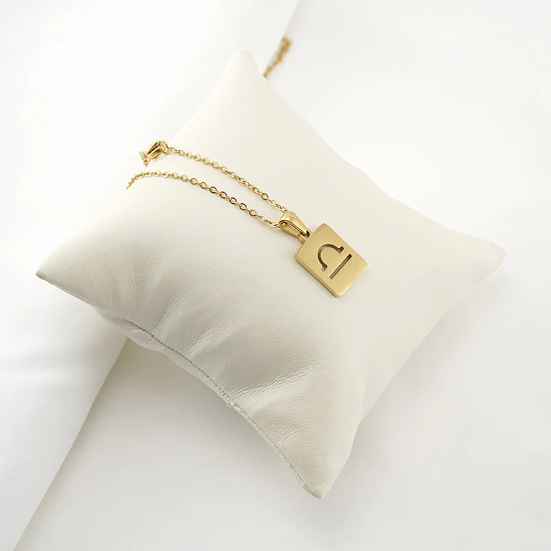 A gold necklace with a dainty chain and rectangular pendant in the  Libra style. The pendant is draped over a white pillow. 