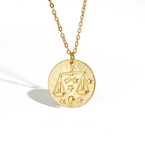 A gold necklace with a dainty chain. The necklace has a coin pendant displaying the astrological sign Libra of the zodiac.