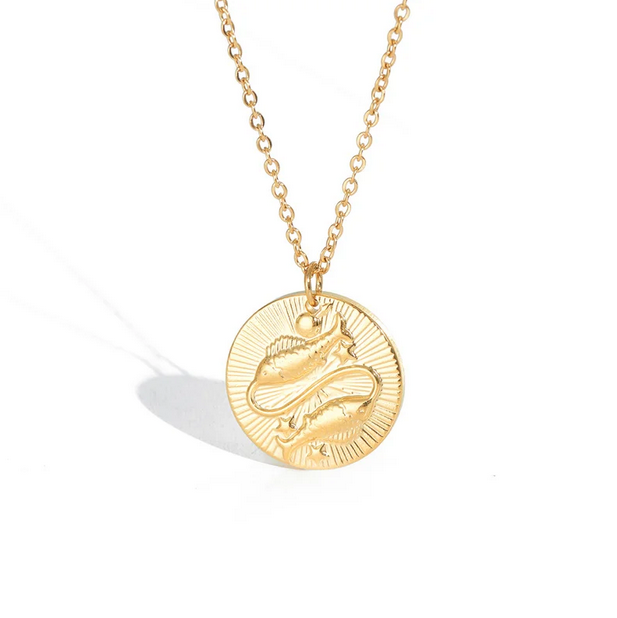 A gold necklace with a dainty chain. The necklace has a coin pendant displaying the astrological sign Pisces of the zodiac.